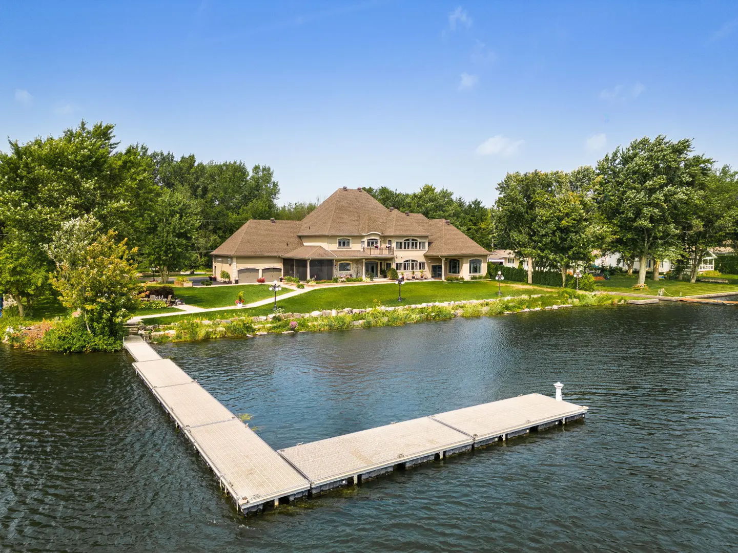 A tan waterfront house with a brown roof sits on a green lawn with a dock extending into the water.