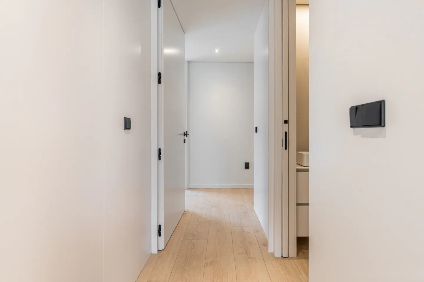 A bright hallway with light wood floors, white walls, and doors leading to a minimalist room and a modern bathroom.