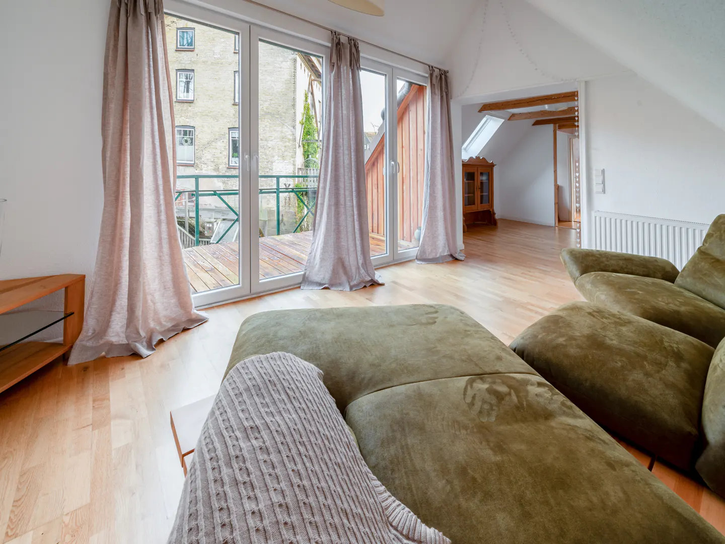 Living room with a green sofa, wood floors, and a balcony with glass doors and light pink curtains.
