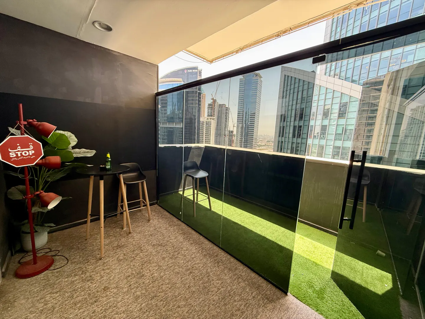 Balcony with city view, glass walls, and artificial turf. A small table, chairs, and a red lamp with a "STOP" sign are visible.