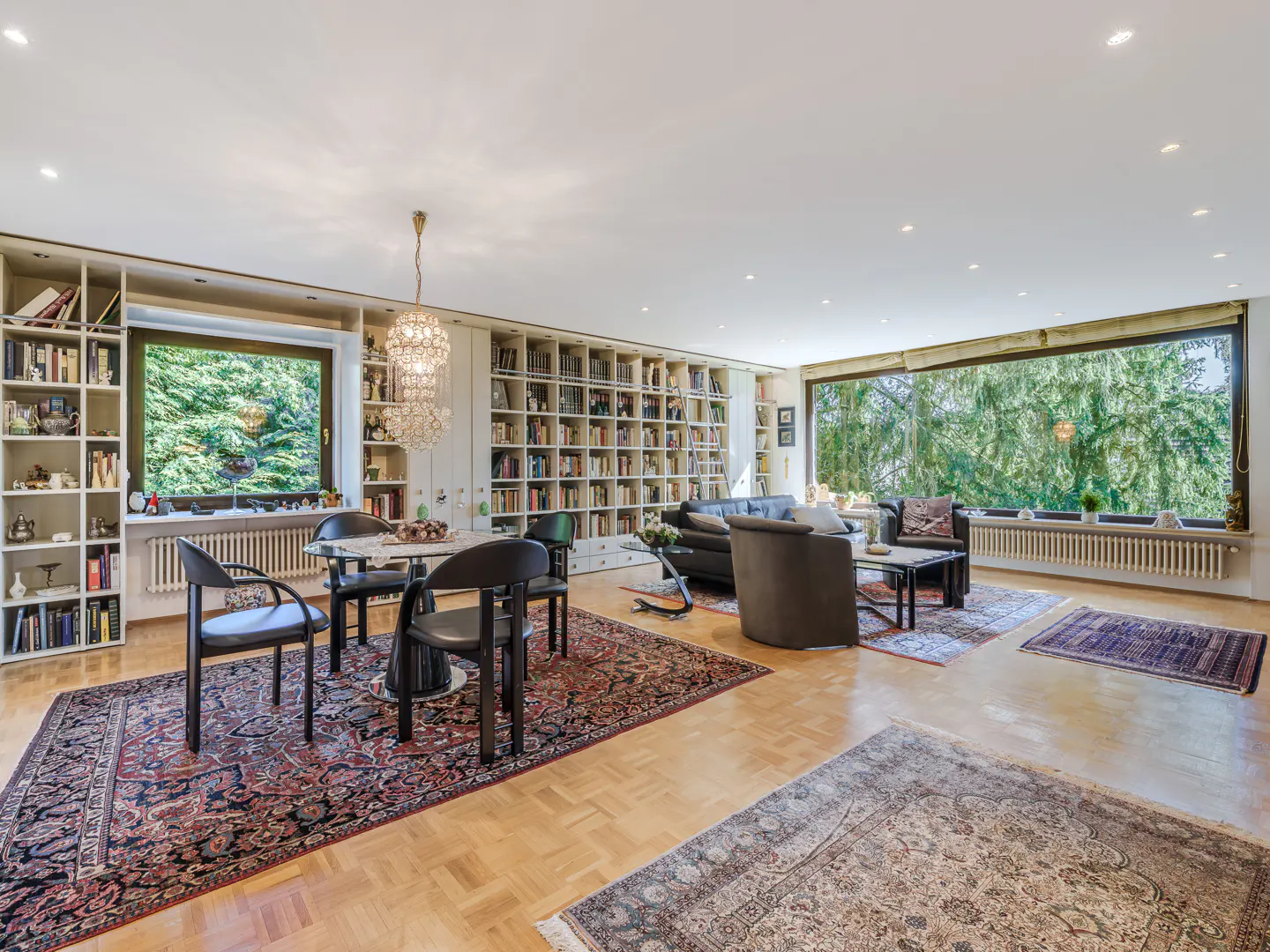 Bright living room with parquet floors, rugs, and floor-to-ceiling bookshelves. A round table with black chairs sits near a large window.