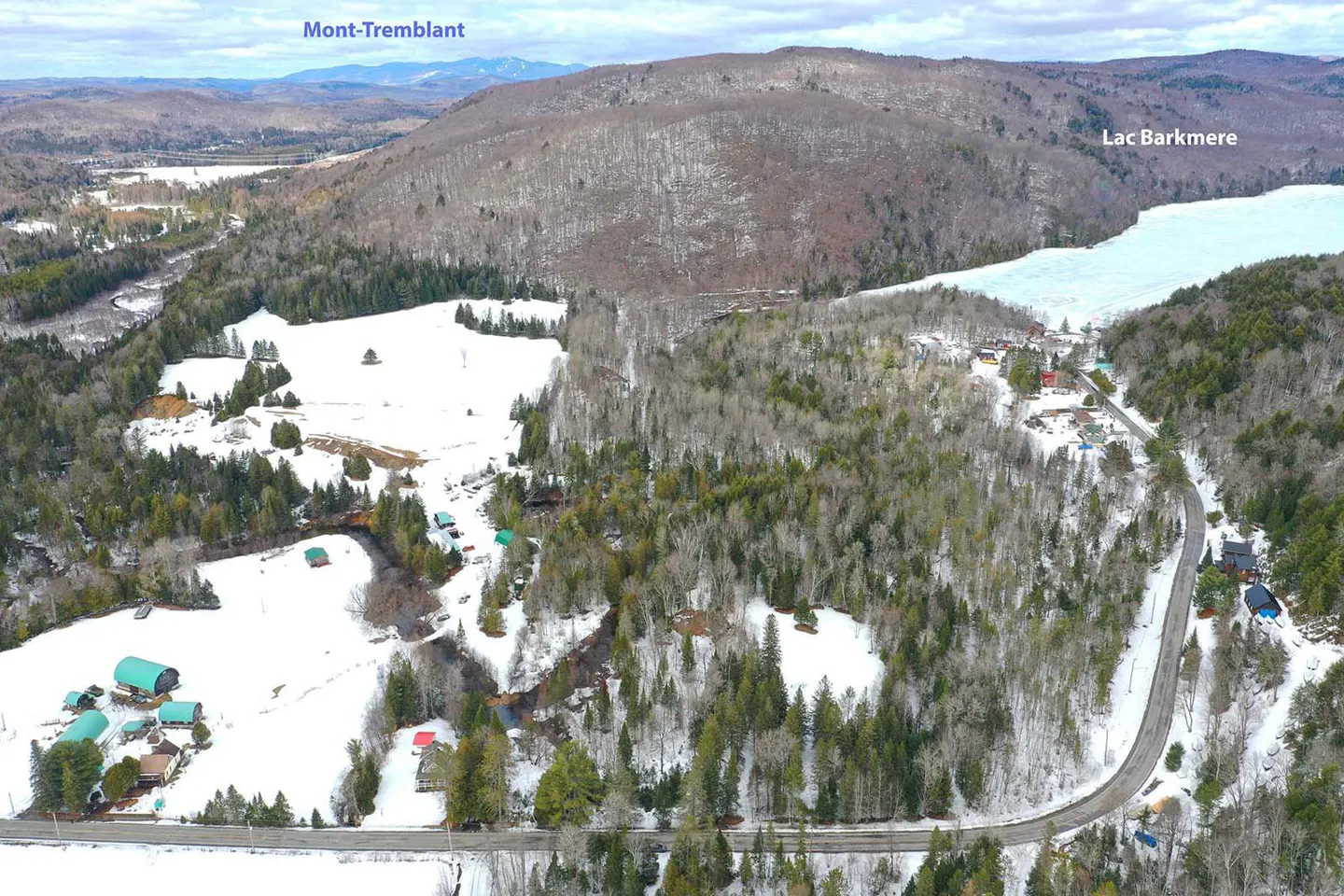 Aerial view of a snowy landscape with Mont-Tremblant in the background and Lac Barkmere on the right. A road winds through the trees and snow-covered fields. Houses are scattered throughout the scene.