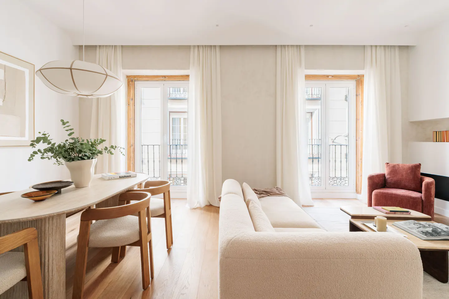 Bright living room with a cream sofa, wood dining table with chairs, and a red armchair. Two windows with white curtains let in natural light.