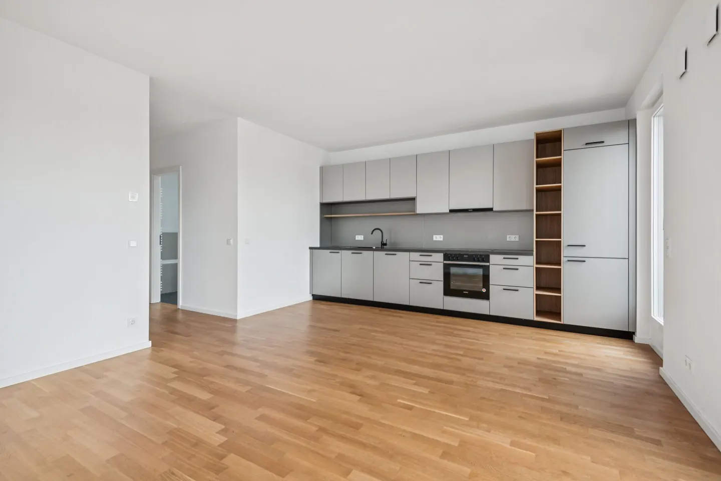 Bright, empty kitchen with light wood floors, white walls, and gray cabinets. A black faucet and oven are visible.