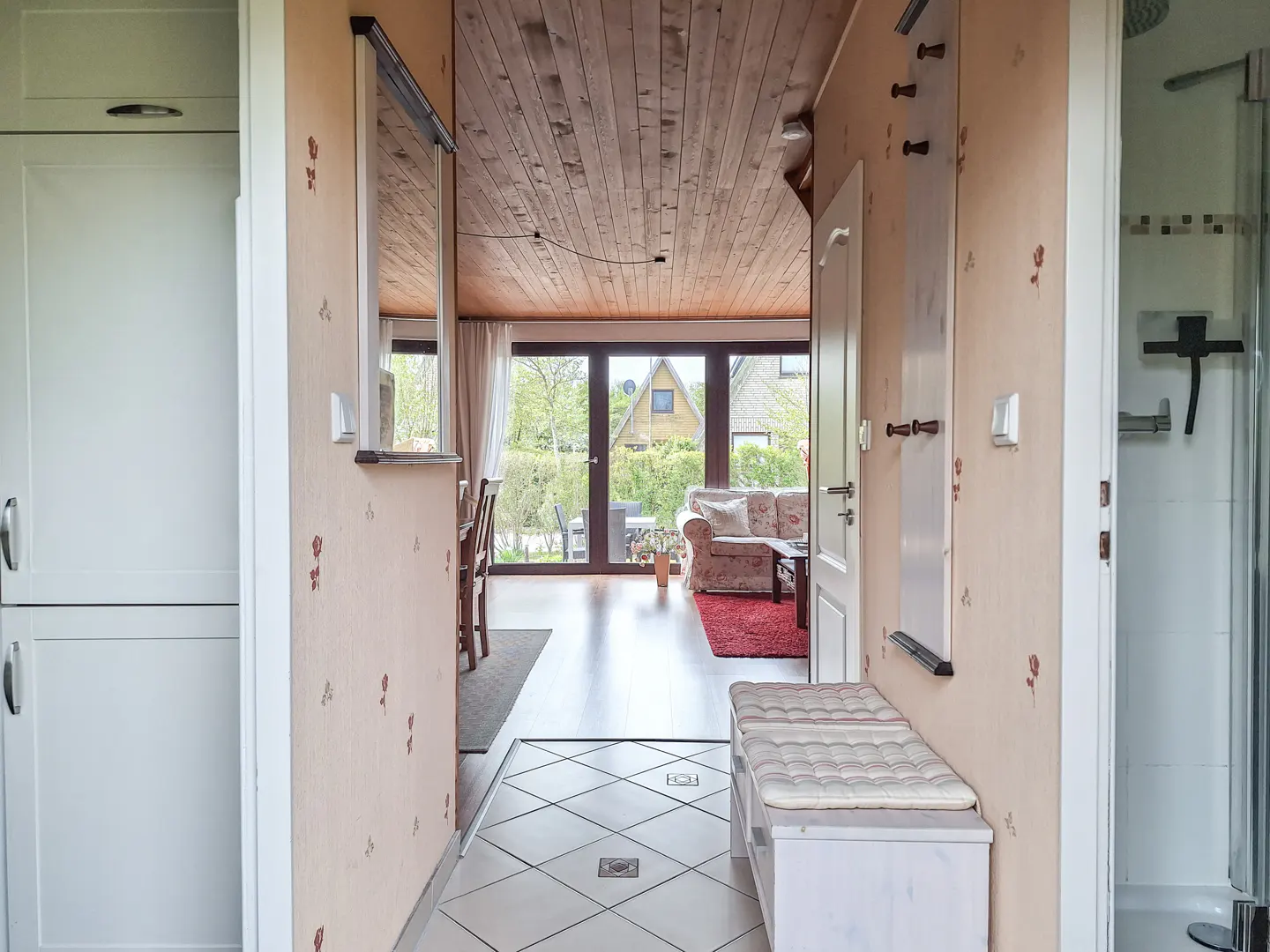Hallway view into a living room with a sofa, red rug, and backyard through glass doors. A white bench sits near a shower room.