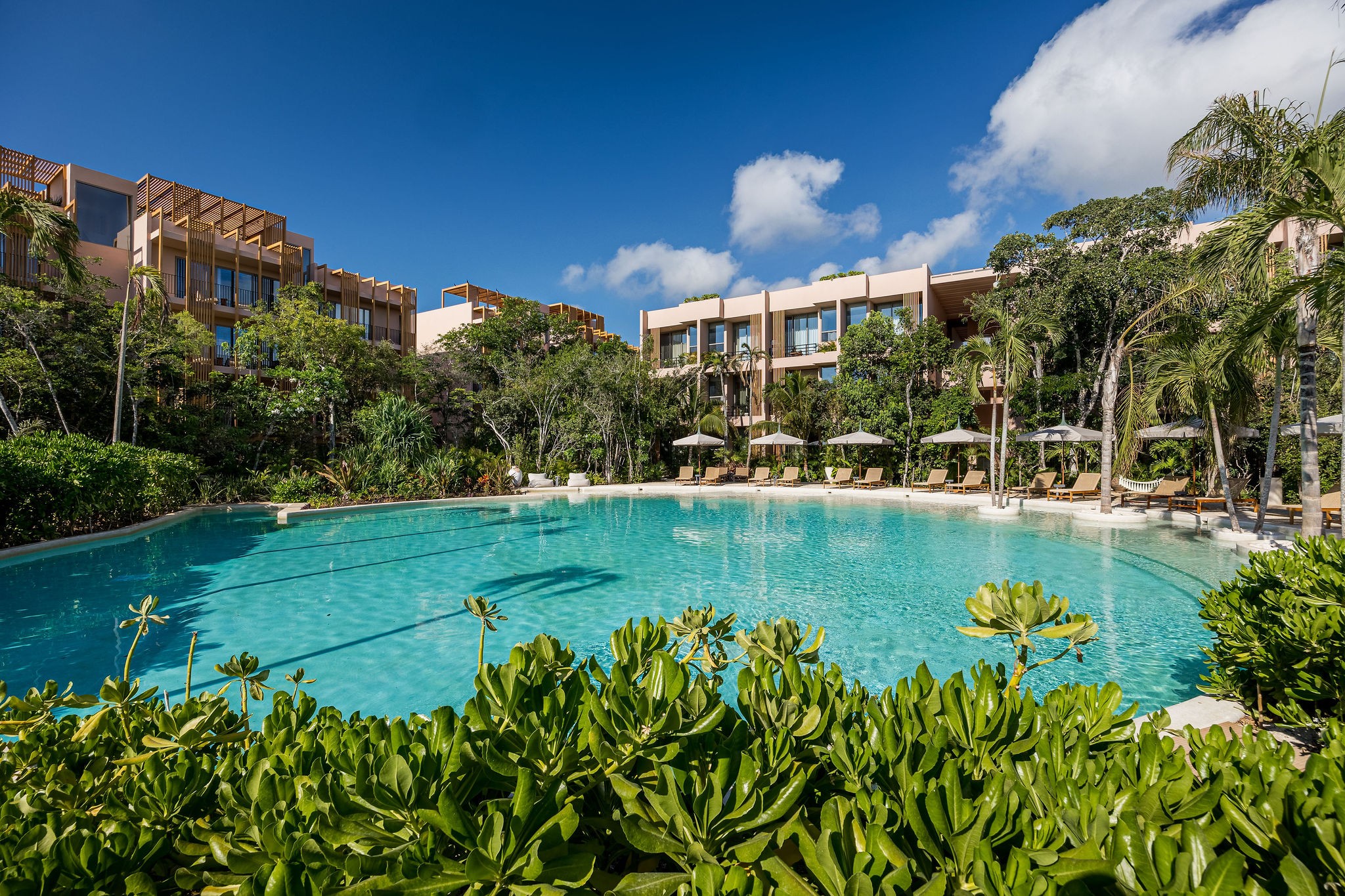 A turquoise swimming pool surrounded by lush tropical greenery, lounge chairs, and modern multi-story buildings.
