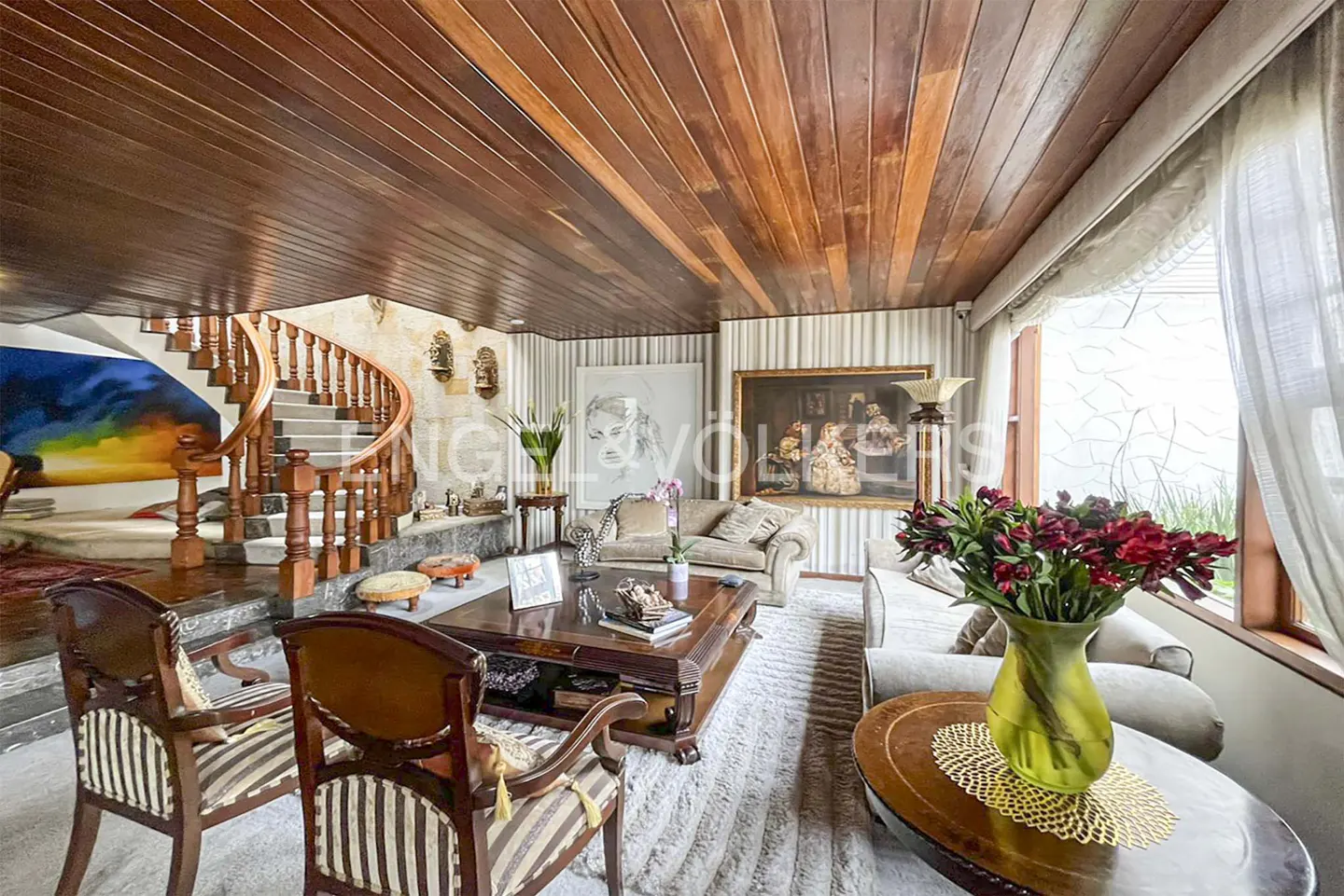 Living room with wood ceiling, spiral staircase, striped chairs, and large paintings. A vase of red flowers sits on a table.