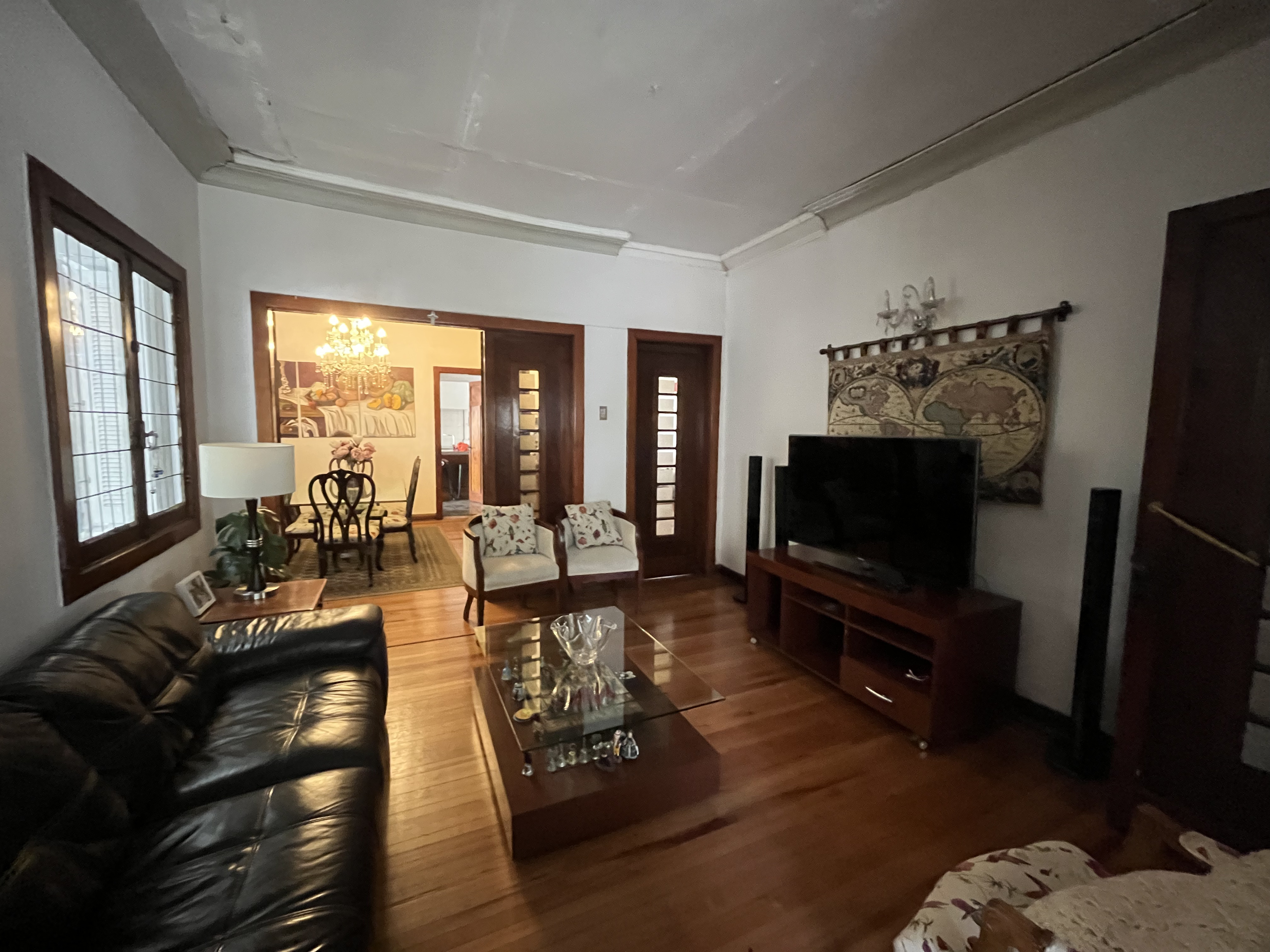 Living room with hardwood floors, black leather sofa, glass coffee table, TV, and dining room visible in the background.