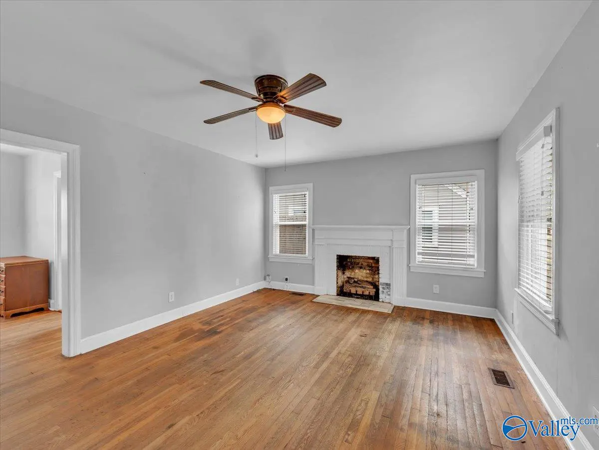 A living room with gray walls, hardwood floors, a white fireplace, and a ceiling fan. Two windows with blinds are visible.