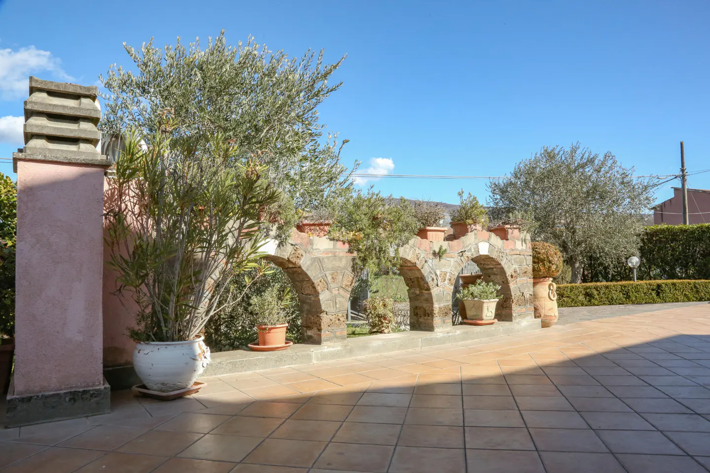 Outdoor patio with stone arches, potted plants, and tiled floor under a blue sky.