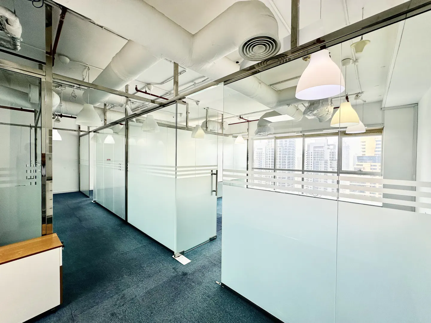 Bright office space with blue carpet, glass-walled cubicles, and white ceilings with exposed ductwork and pendant lights.