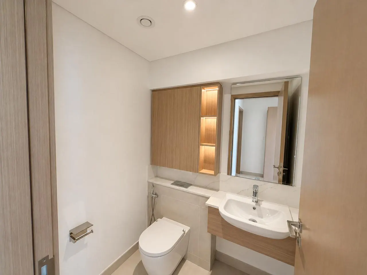 A bright bathroom with a white toilet, sink, and wood cabinet. A mirror reflects the doorway.