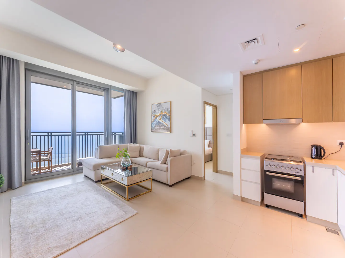 Bright apartment with ocean view. Beige sectional sofa, mirrored coffee table, and balcony with seating. Modern kitchen with wood cabinets.
