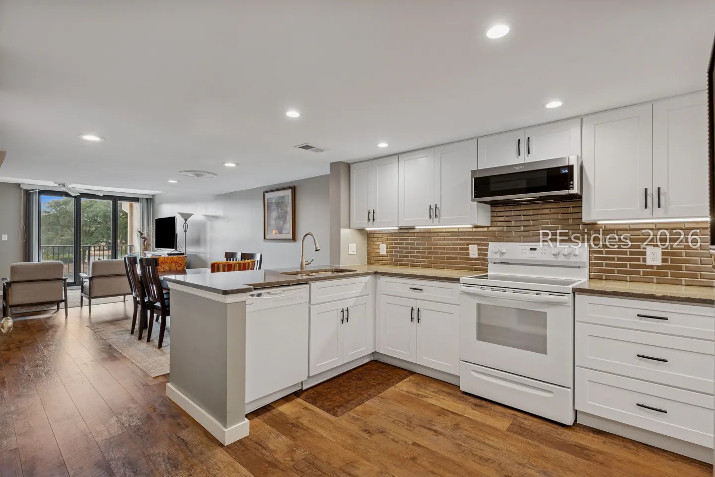 Open concept kitchen with white cabinets, stainless steel appliances, and wood floors. Living room with balcony in background.