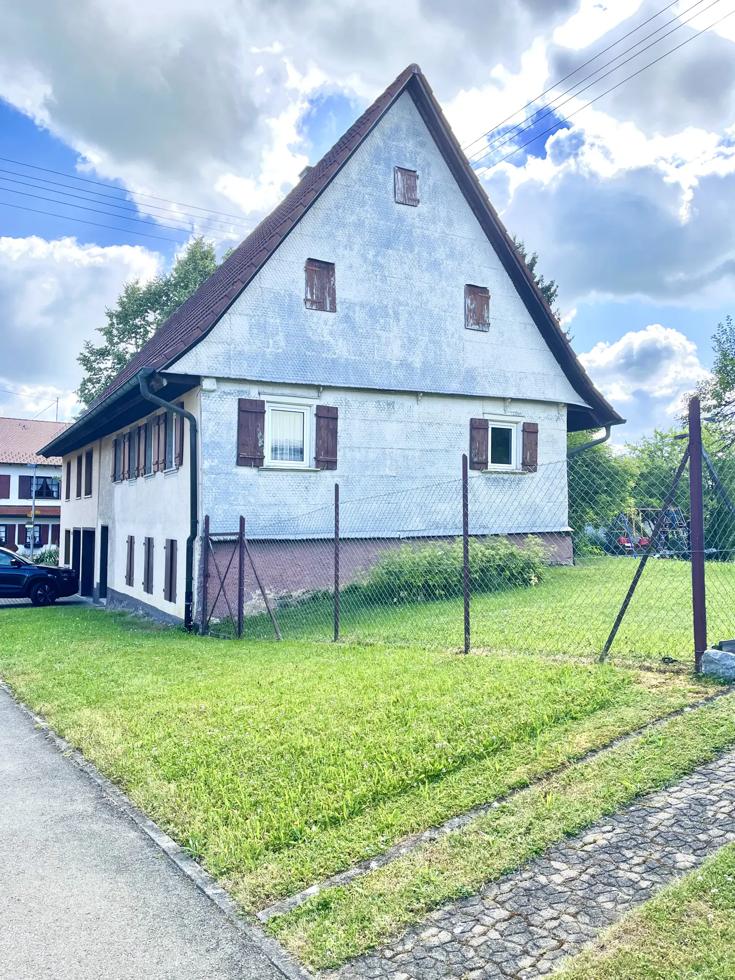 A two-story white house with brown shutters and a brown roof, surrounded by a green lawn and a chain-link fence.