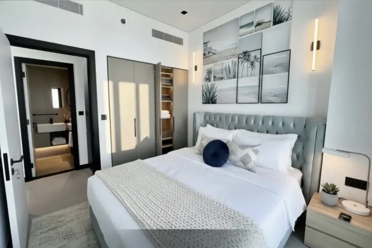 A modern bedroom with a gray tufted headboard, white bedding, and beach-themed art. A bathroom is visible through an open door.