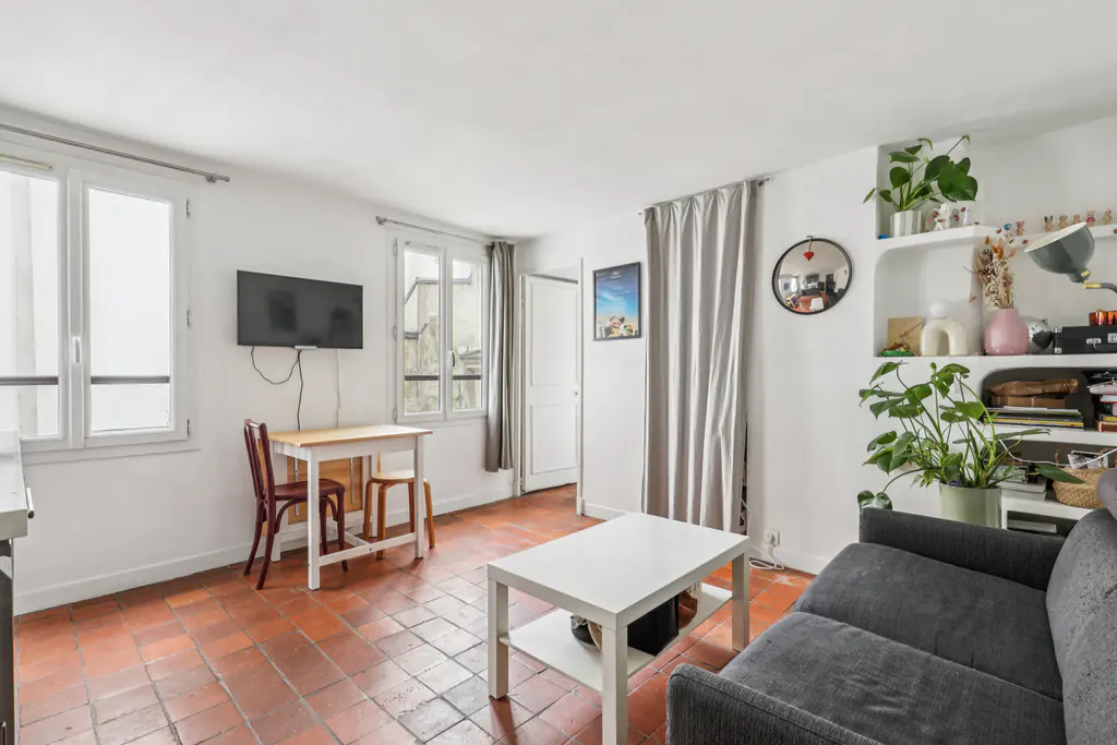 Bright living room with terracotta tile floor, white walls, grey sofa, white coffee table, dining table, and shelves with plants.