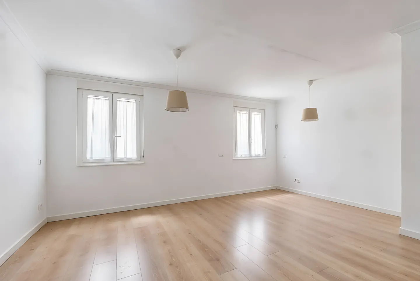 Bright, empty room with light wood floors, white walls, two windows with sheer curtains, and two pendant lights.