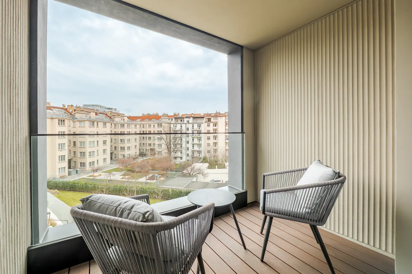 Modern balcony with two grey woven chairs, a small table, and a view of urban apartment buildings and a courtyard.
