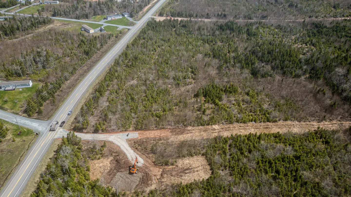 Aerial view of a wooded lot with an orange excavator clearing land near a road intersection.