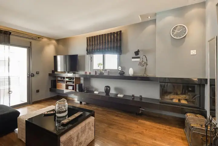 Living room with hardwood floors, gray walls, and a black entertainment center with a fireplace. A black coffee table sits on a beige ottoman.