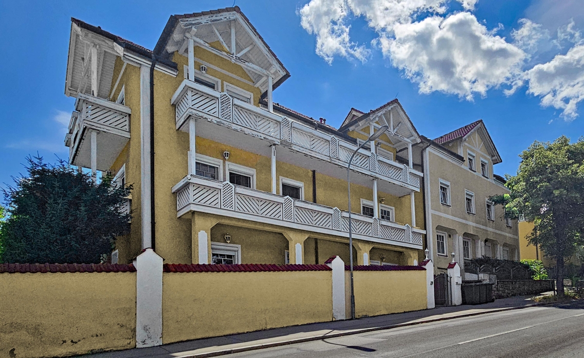 A yellow multi-story building with white balconies and a matching perimeter wall under a bright blue sky with clouds.