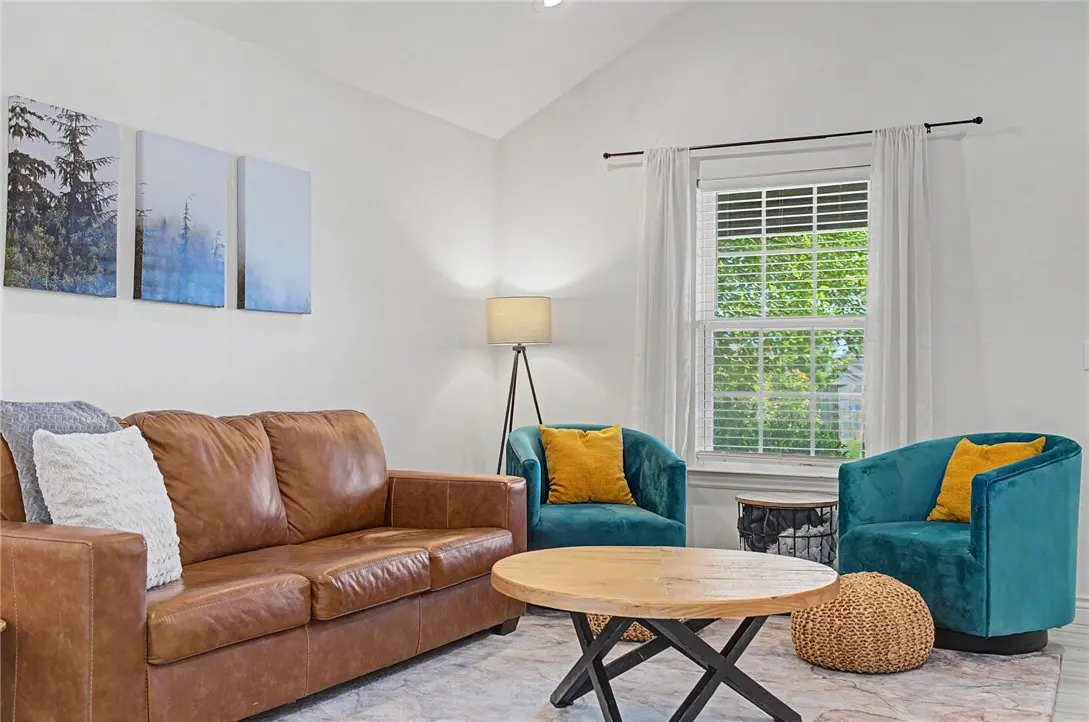 Living room with brown leather sofa, teal chairs with yellow pillows, and round wood coffee table. Forest art on the white wall.