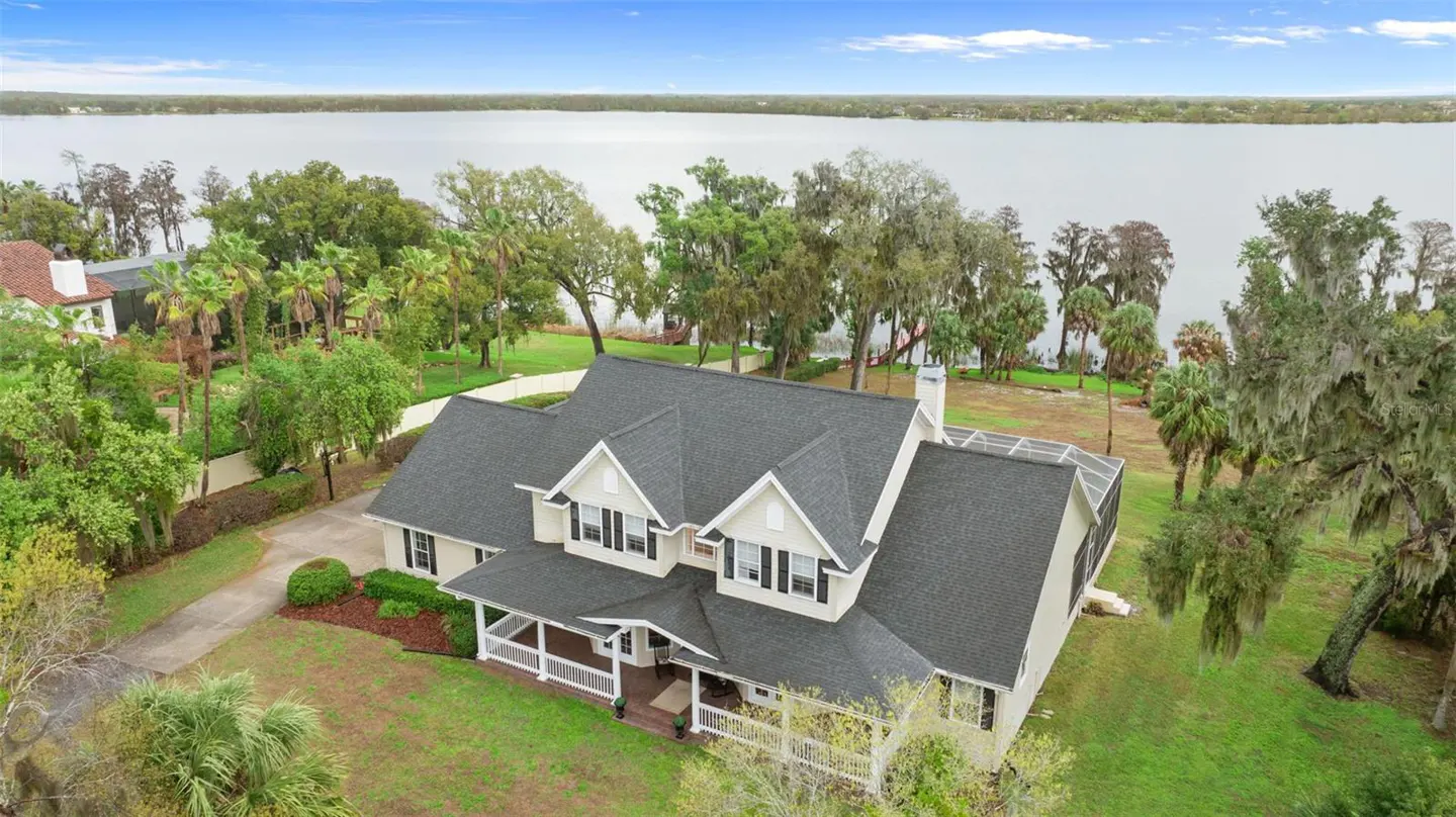Aerial view of a two-story beige house with a gray roof, white trim, and a front porch, set against a backdrop of a lake and trees.