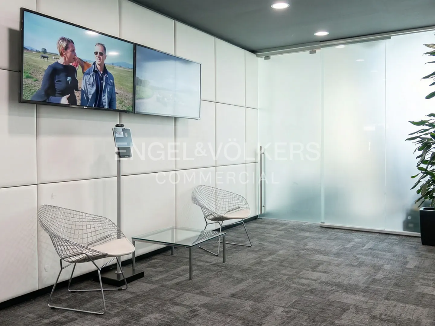 Waiting area with two wire chairs, glass table, and two TVs on a white paneled wall. Engel & Völkers logo on frosted glass door.