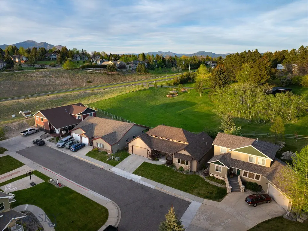 Aerial view of a suburban street with houses, cars, green lawns, and mountains in the background.