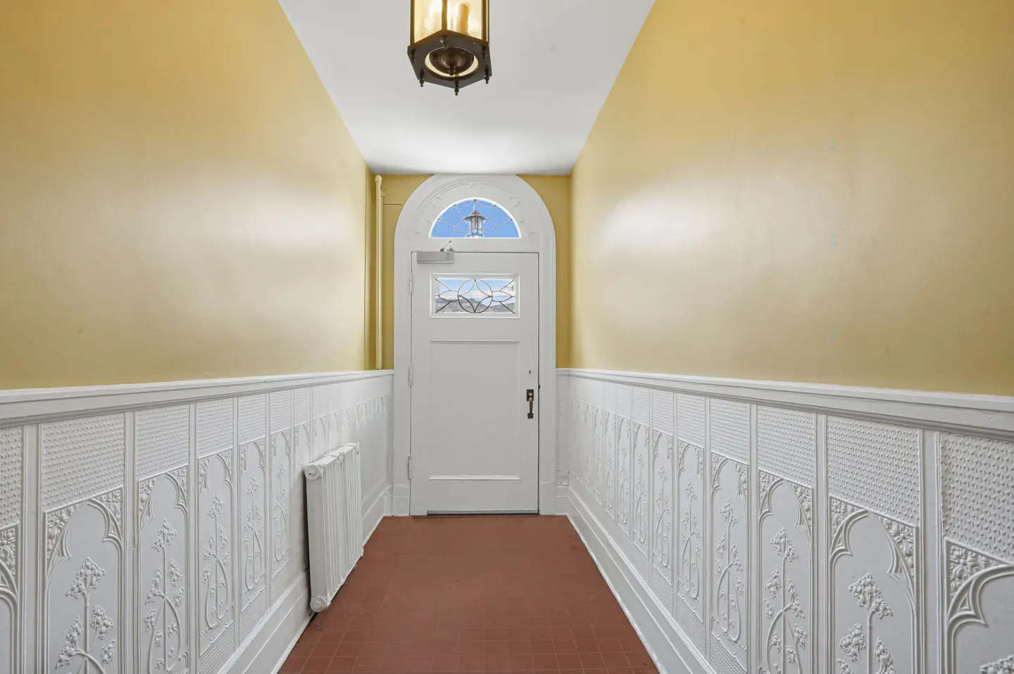 Hallway with yellow walls, white wainscoting, and a white door with a window. A brown lantern hangs from the ceiling.