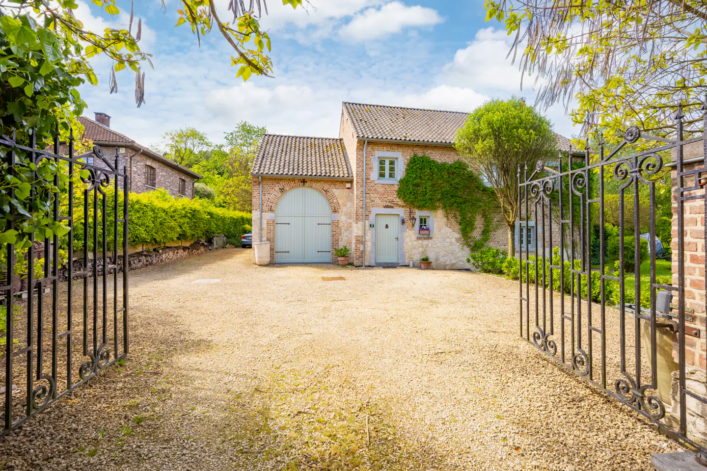 Exterior view of a stone house with a gravel driveway, seen through an ornate black metal gate on a sunny day.