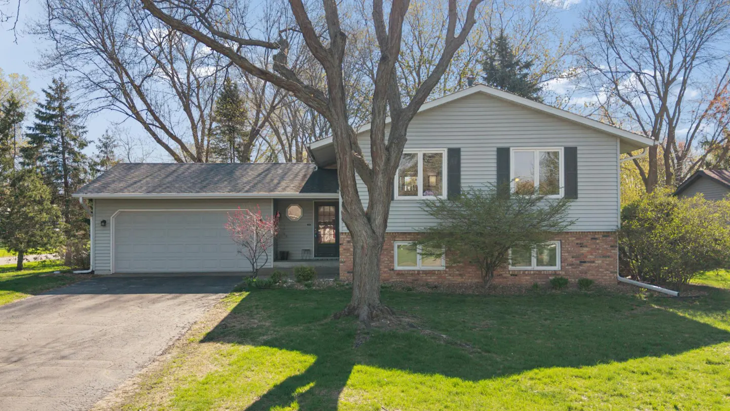 Exterior view of a two-story, light gray house with a brick foundation and a two-car garage. A large tree is in the front yard.