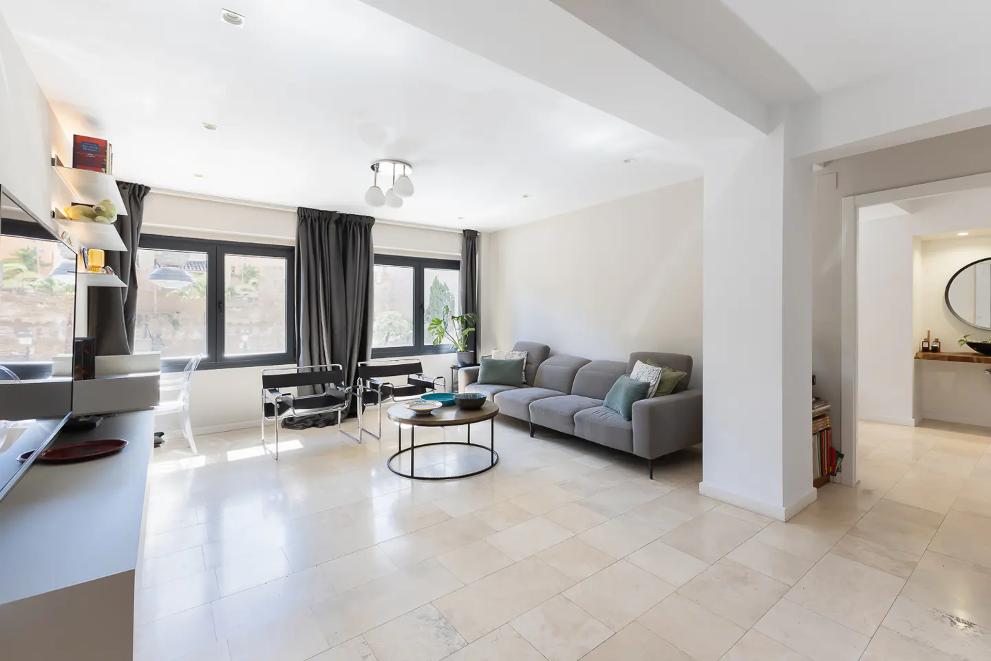 Bright living room with a gray sofa, black chairs, and a round coffee table on a beige tiled floor. Gray curtains frame the windows.
