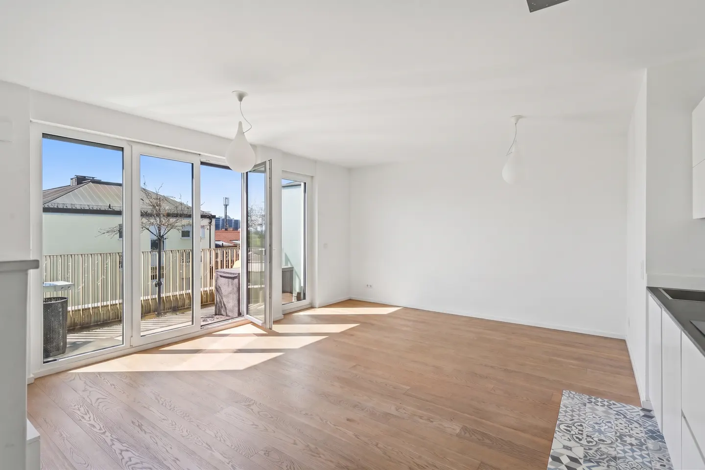 Bright, modern apartment with hardwood floors, white walls, and large glass doors leading to a balcony. Two pendant lights hang from the ceiling.
