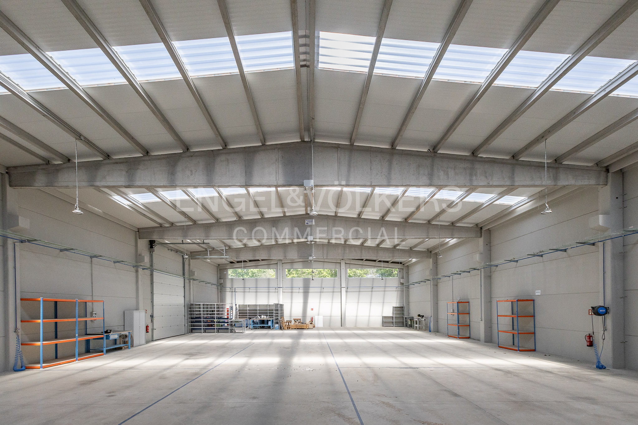 Spacious, empty industrial warehouse with high ceilings, skylights, concrete floors, and metal shelving units along the walls.