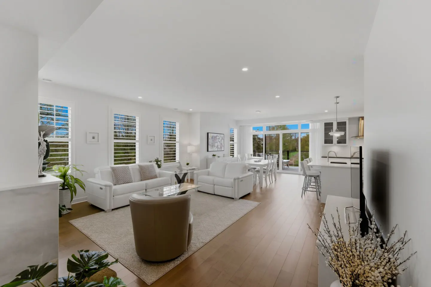 Bright, open-concept living space with white sofas, a tan chair, and a dining area leading to a patio with a green lawn.
