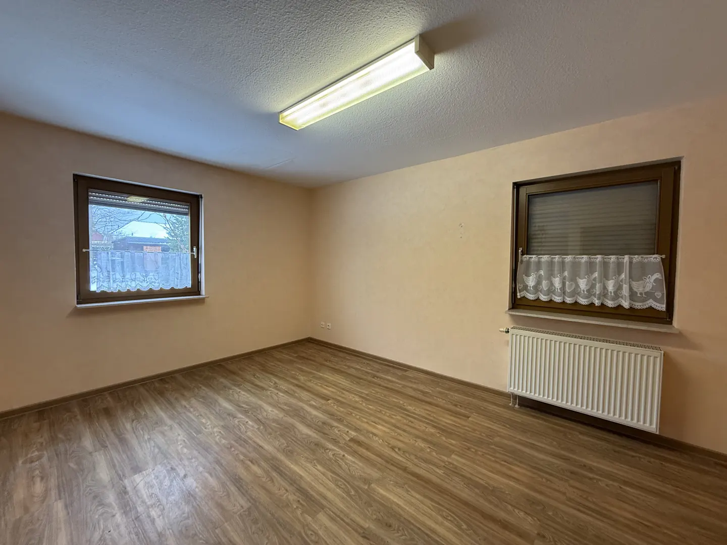 Empty room with wood-look floors, peach walls, and two windows. One window has a white lace curtain. A white radiator sits below the window.
