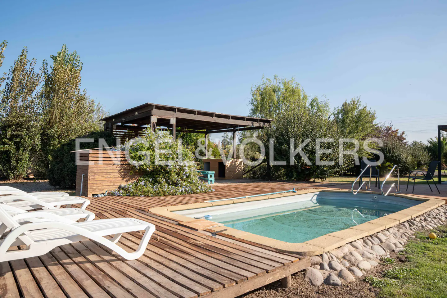 Outdoor pool area with wooden deck, lounge chairs, and a pergola under a clear blue sky.