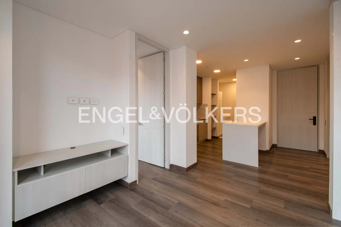 An interior view of a modern apartment with light wood floors, white walls, and recessed lighting. A white media console sits below the Engel & Völkers logo.