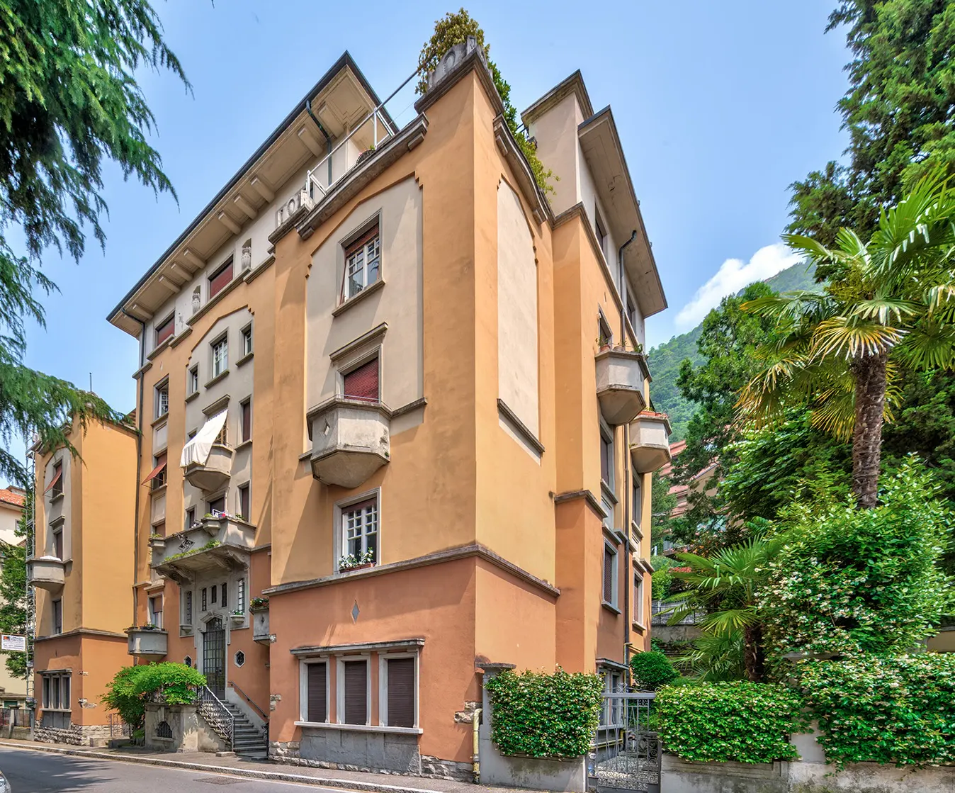 A multi-story, peach-colored apartment building with balconies and lush greenery on a sunny day.