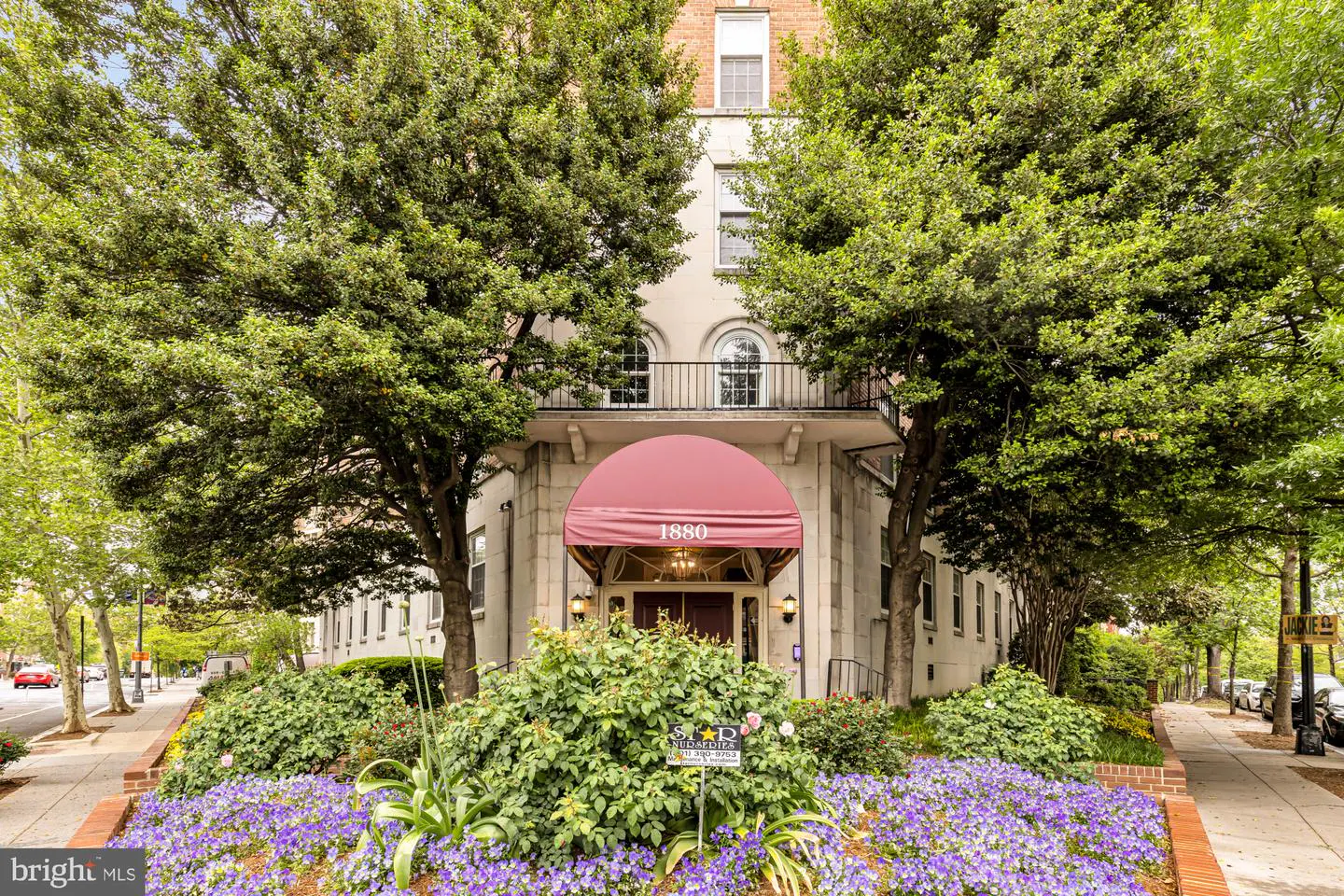 Exterior of a light-colored building with a red awning over the entrance, number 1880, framed by green trees and purple flowers.