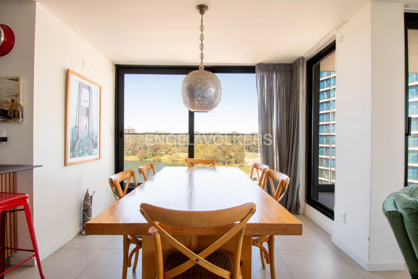 Bright dining room with a long wooden table and chairs, a globe pendant light, and a large window with a view of a lake and trees.