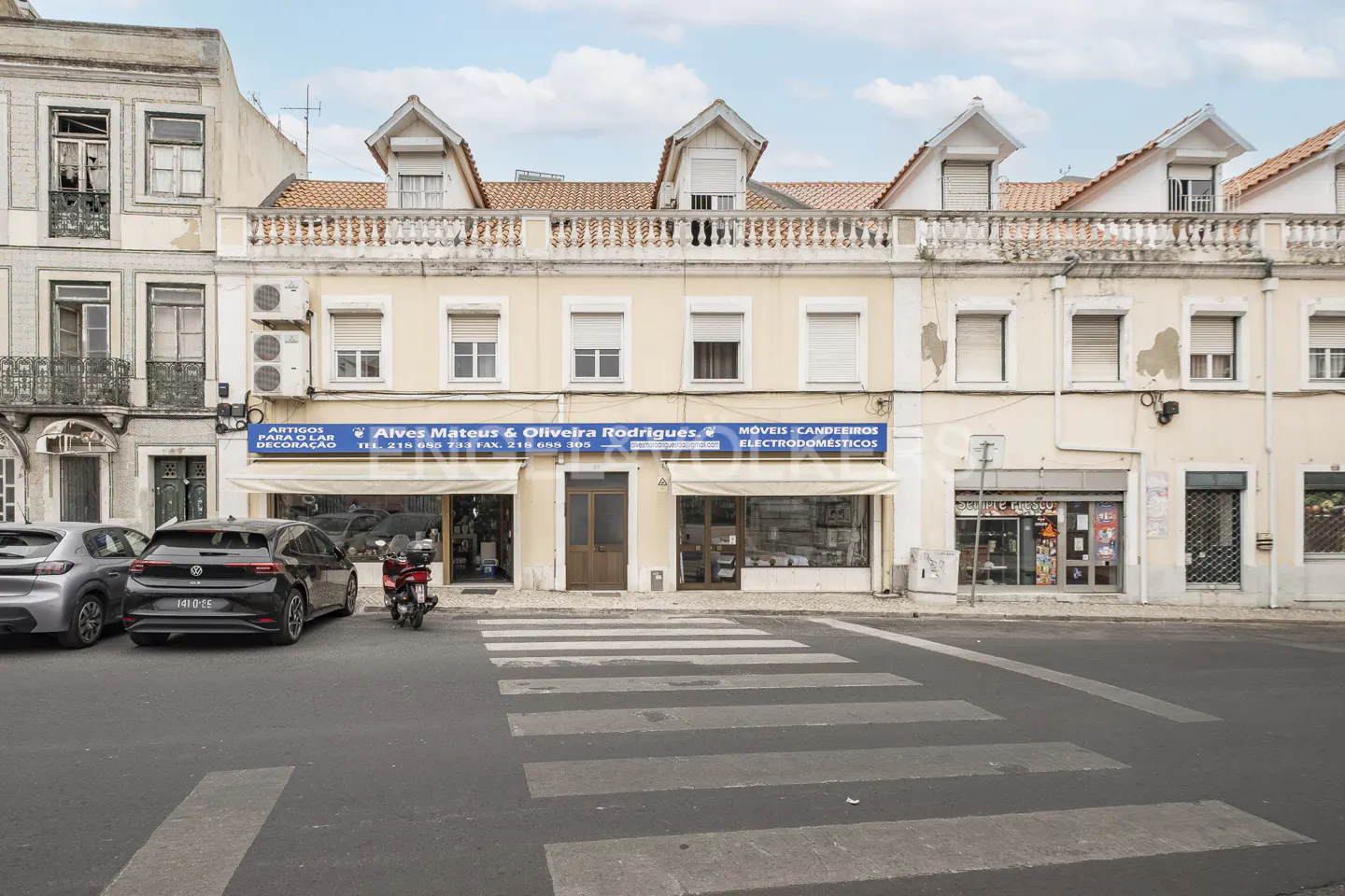 Street view of a two-story building with shops on the ground floor, cars parked on the street, and a crosswalk in front.