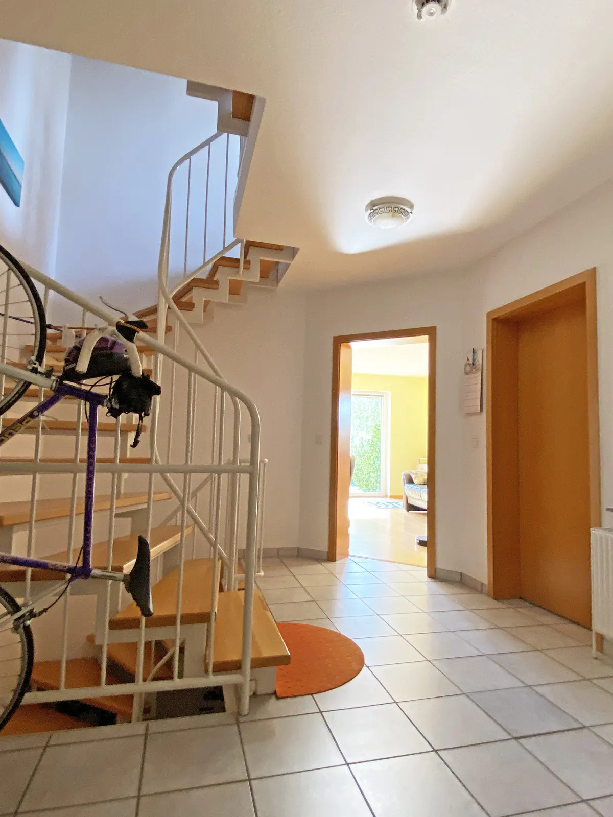 Bright foyer with white walls, tiled floor, and a winding staircase with a bicycle leaning against it. Two doorways lead to other rooms.
