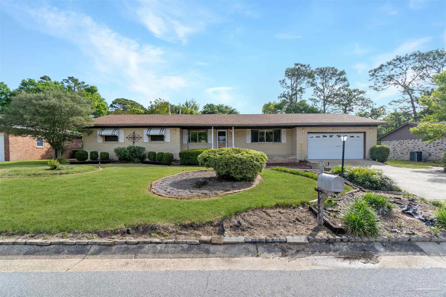 Beige ranch-style house with brown roof, white trim, and green lawn under a blue sky. A circular flower bed is in the front yard.