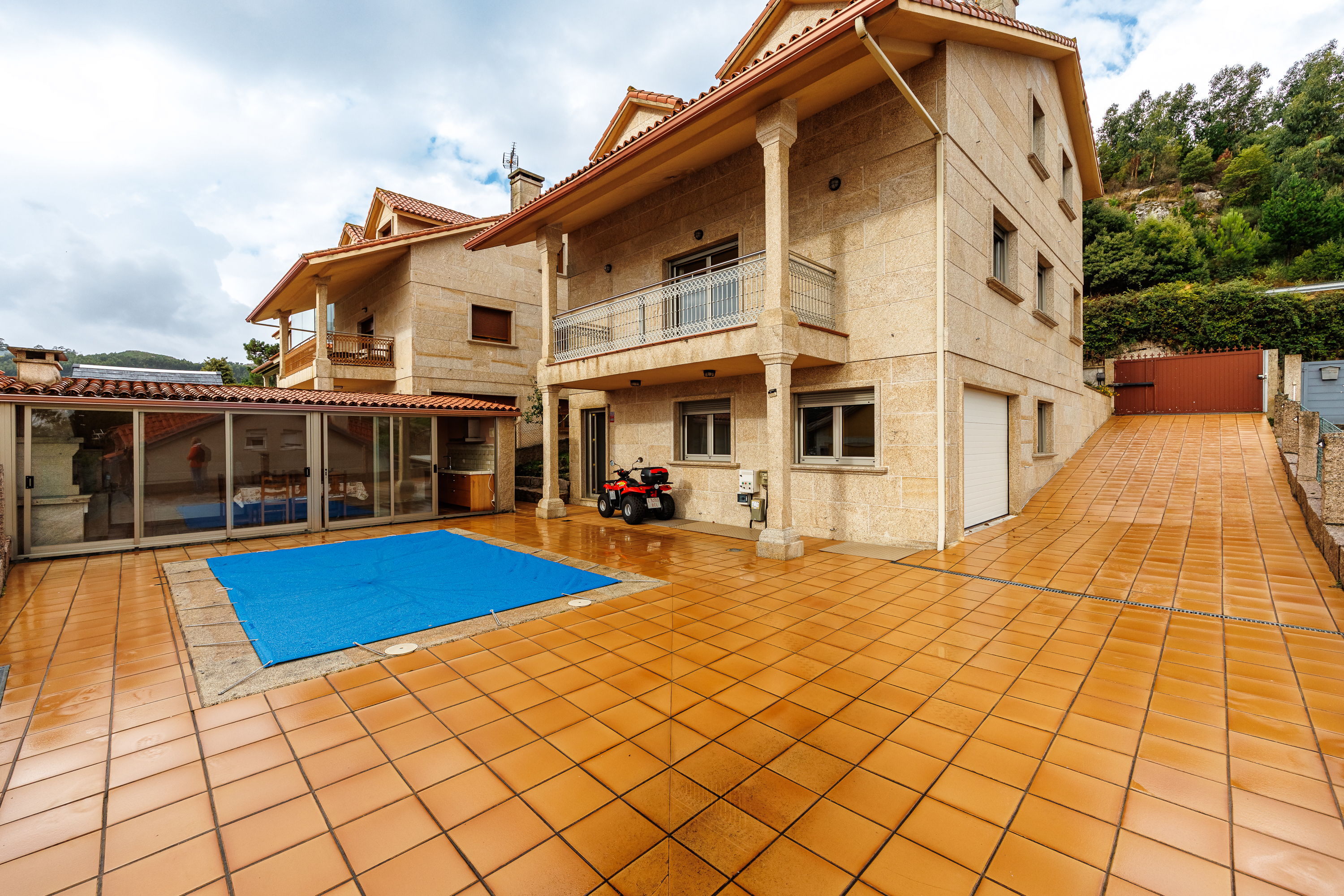 Two-story stone house with a tiled patio, covered pool, and a red ATV parked near the entrance under a balcony. [AI-generated text]