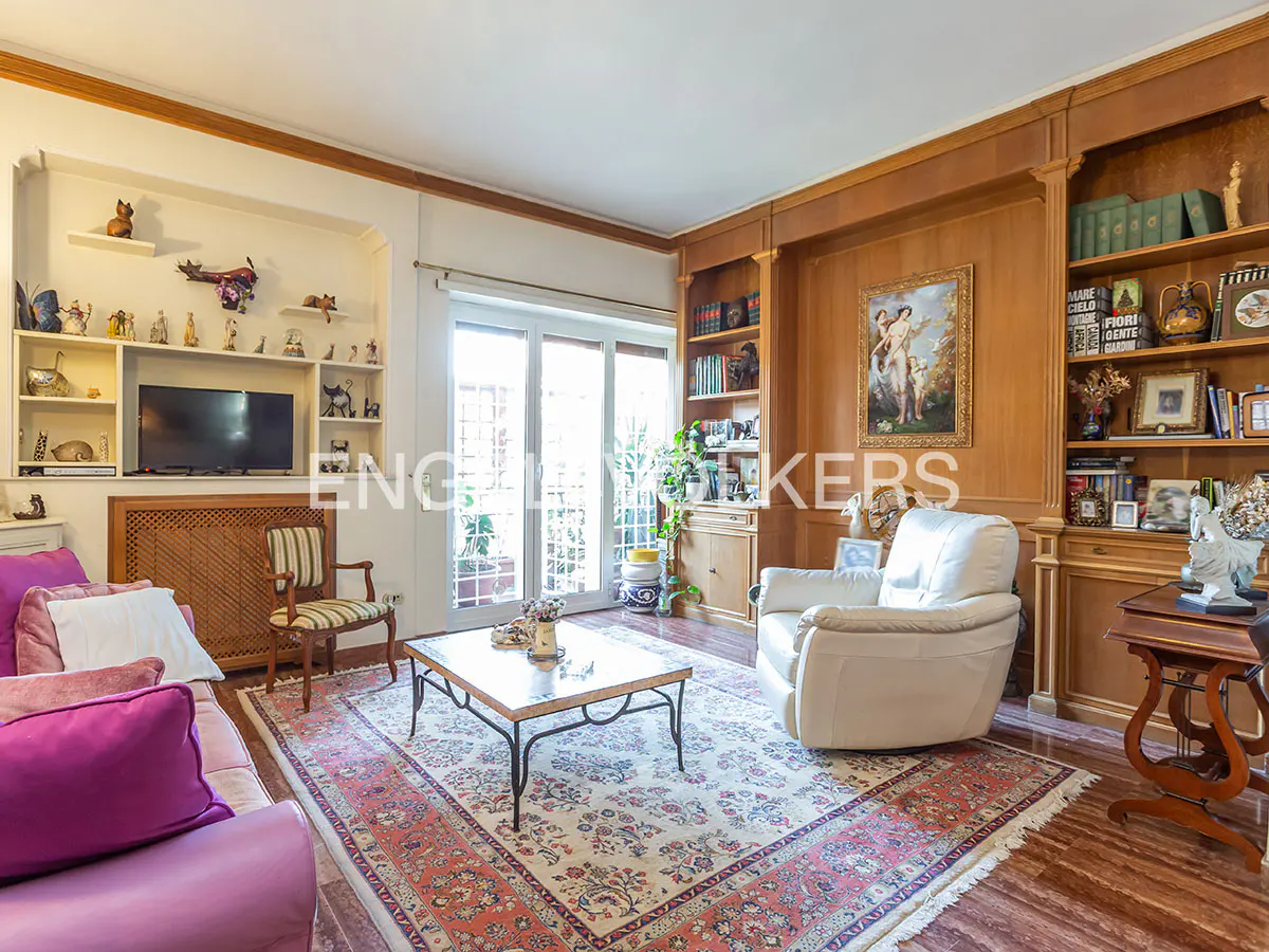 Living room with a pink sofa, white armchair, and a patterned rug. Shelves display decorative items, and a painting hangs on the wood-paneled wall.