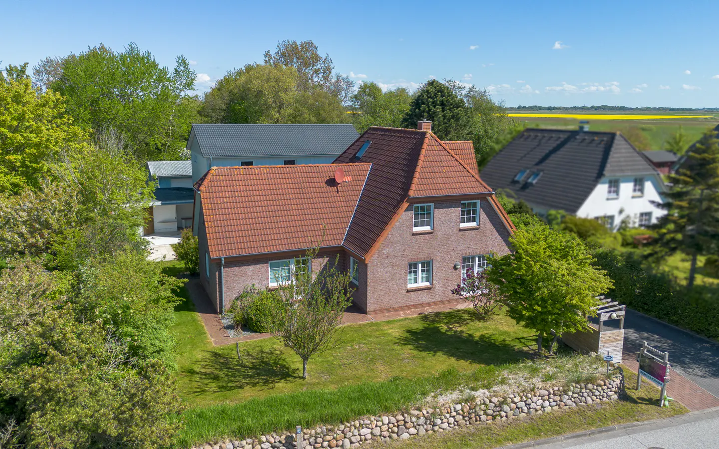Aerial view of a brick house with a red tile roof, surrounded by green trees and a lawn. A stone wall borders the property.