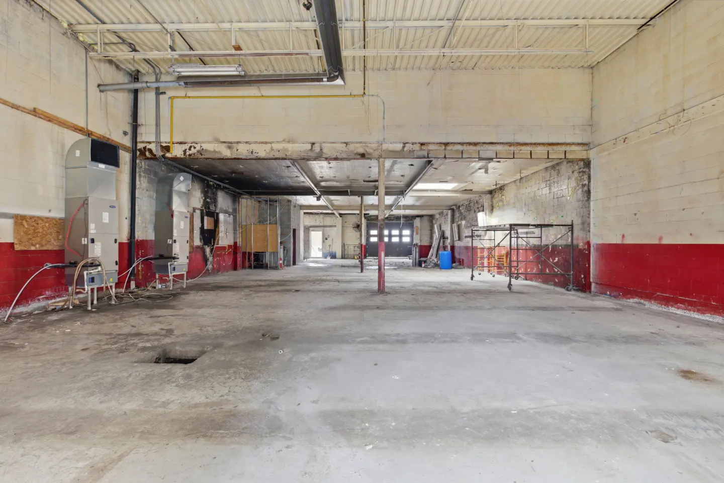 Large, empty commercial space with concrete floor, red trim, and overhead pipes. A garage door is visible in the background.